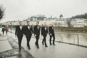 Fünf Männer im Smoking schlendern an einem Fluss entlang in einer europäischen Stadt. Im Hintergrund thront ein Schloss auf einem Hügel. Der bewölkte Himmel verleiht dem Ganzen eine dramatische Note und schafft die perfekte Kulisse für alle, die von einer eleganten Hochzeit träumen. Charmante Gebäude säumen das gegenüberliegende Flussufer.