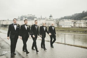 Fünf Männer im Smoking schlendern einen Uferweg entlang, die historische Stadt und das Schloss bilden eine malerische Kulisse. Es ist ein bewölkter Tag, perfekt zum Heiraten, denn die Gruppe scheint in Gespräche und Gelächter vertieft zu sein und bespricht vielleicht letzte Details ihrer Hochzeitsplanung.