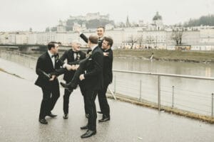 Fünf Männer in Anzügen lachen und heben einen ihrer Freunde hoch, vielleicht feiern sie eine Hochzeit an einer Uferpromenade. Im Hintergrund ist eine Stadtlandschaft mit historischen Gebäuden und einem Schloss auf einem Hügel zu sehen, was zur freudigen Atmosphäre dieses unvergesslichen Hochzeitsmoments beiträgt.