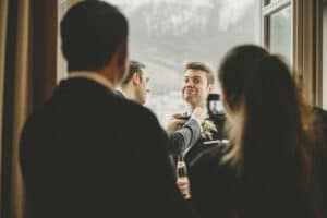 Ein Bräutigam im Anzug steht lächelnd neben einem Fenster und genießt die Freude der Hochzeit, während eine andere Person seine Ansteckblume zurechtrückt. Zwei weitere sind ihm zugewandt, einer hält ein Telefon in der Hand, um den Moment festzuhalten. Draußen tragen Bäume und eine verschneite Landschaft zu dieser malerischen Hochzeitsszene bei.