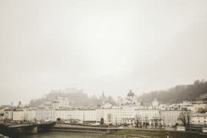 Nebeliger Blick auf eine europäische Stadt mit historischen Gebäuden entlang eines Flussufers. Die Skyline, perfekt für eine romantische Hochzeit, zeigt ein großes Schloss auf einem Hügel, mit im Nebel sichtbaren Kirchenkuppeln, was eine ruhige und stimmungsvolle Szenerie schafft, die ideal für Hochzeiten ist.