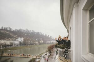 Vier Menschen in Anzügen stehen auf einem Balkon und stoßen mit Flaschen auf ihre Hochzeit an. Vom Balkon aus hat man einen Blick auf einen Fluss und eine malerische Landschaft mit Gebäuden am Hang unter einem wolkigen Himmel. Fußgänger schlendern unten am Flussufer entlang und bemerken die fröhliche Hochzeitsplanung oben nicht.