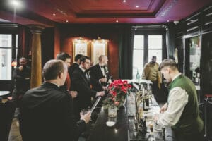 Ein Barkeeper in weißem Hemd und grüner Weste serviert Getränke an eine Gruppe von Männern in Anzügen, die sich vielleicht über Heiraten unterhalten. Der schwach beleuchtete Raum hat rote Wände und die Theke ist mit roten Weihnachtssternen geschmückt. An der Wand hängen mehrere gerahmte Bilder.