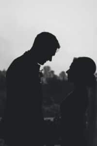 Silhouette eines Paares, das sich vor einem verschwommenen Hintergrund aus Bäumen und Himmel gegenübersteht. Die Figuren scheinen sich an den Händen zu halten, was ein Gefühl von Intimität und Verbundenheit vermittelt und an die zärtlichen Momente in Hochzeitsplanung erinnert.