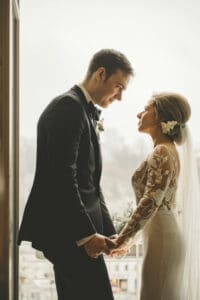 Ein Paar im Hochzeitskleid steht einander gegenüber und hält sich an den Händen. Der Bräutigam trägt einen schwarzen Anzug und die Braut ein weißes Kleid mit Spitzenärmeln und Schleier. Sie stehen drinnen, in der Nähe eines Fensters, das von sanftem, natürlichem Licht erhellt wird – ein perfekter Moment auf ihrer Hochzeitsreise.