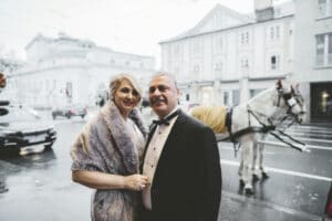 Ein Paar in festlicher Kleidung steht auf einer Stadtstraße und plant vielleicht seine Hochzeit. Die Frau trägt einen Pelzschal und der Mann einen Smoking. Hinter ihnen glitzert eine Pferdekutsche auf dem nassen Asphalt, mit Stadtgebäuden im Hintergrund.