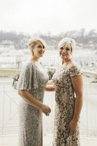 Zwei Frauen in eleganten, funkelnden Kleidern stehen auf einem Balkon mit einer malerischen Kulisse aus nebliger Stadtlandschaft und Hügeln im Hintergrund. Sie lächeln in die Kamera und genießen die Aussicht und die Gesellschaft der anderen – ein perfekter Moment, um jede Hochzeitsplanung zu inspirieren.