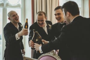 Fünf Männer in formellen Anzügen stoßen fröhlich mit Bierflaschen an, um einen besonderen Moment ihrer Hochzeitsplanung auszusprechen. Sie stehen in einem warm beleuchteten Raum mit einem großen Fenster und eleganter Dekoration im Hintergrund.