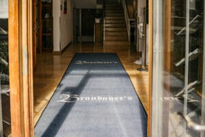 Eingangsflur mit blauem Teppich, der zu einer Treppe führt, perfekt für die Hochzeitsplanung. Der Teppich zeigt elegant „Brandauer’s“ in Weiß. Sonnenlicht wirft bezaubernde Schatten auf den Boden, während Holztüren den Blick einrahmen und eine romantische Bühne für Hochzeitsträume schaffen.