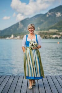 Eine Frau steht auf einem Holzsteg an einem See, umgeben von Bergen, und träumt von ihrer Hochzeit. Sie trägt ein traditionelles Kleid mit einer blau-grünen Schürze und hält einen Strauß weißer und blauer Blumen in der Hand. Der Himmel ist teilweise bewölkt und schafft eine ruhige Kulisse, perfekt für eine Hochzeit.