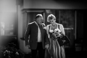 Ein Schwarzweißfoto zeigt ein lächelndes Paar, das Hand in Hand geht und an ihren Hochzeitstag erinnert. Die Frau hält einen Blumenstrauß in der Hand, ihr traditionelles Kleid weht, während der Mann in einer legeren Jacke sie begleitet. Der sanft verschwommene Hintergrund unterstreicht ihre freudigen Ausdrücke in diesem besonderen Moment der Heirat.
