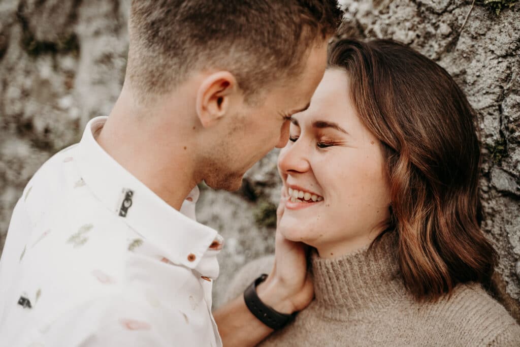 Ein Paar steht dicht beieinander an einer Steinmauer und lächelt sich an seinem Hochzeitstag herzlich an. Die Frau mit schulterlangem braunem Haar trägt einen beigen Pullover, während der Mann kurze Haare hat und ein helles Hemd trägt.
