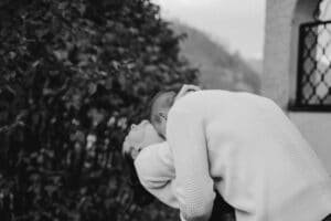 Ein Schwarzweißfoto zeigt ein Paar, das sich in einem zärtlichen Kuss umarmt und die Essenz ihrer Hochzeit verkörpert. Die Person auf der linken Seite lehnt sich zurück, während sich die andere vorbeugt, umgeben von Laub und einer Außenwand mit einem Fenster. Beide tragen leichte Pullover, was ihre zeitlose Liebe widerspiegelt.