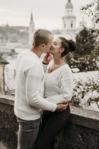 Ein Paar in weißen Pullovern küsst sich romantisch, während es an eine Steinmauer lehnt. Der perfekte Moment inmitten ihrer Hochzeitsplanung. Mit einer verschwommenen Stadtlandschaft und Uhrtürmen im Hintergrund verkörpert die Atmosphäre Gelassenheit, während sie von ihrer bevorstehenden Hochzeit träumen.