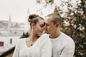 Ein Paar in weißen Pullovern steht eng umschlungen im Freien zwischen Bäumen, im Hintergrund ist eine verschwommene Stadtlandschaft zu sehen. Mit geschlossenen Augen genießen sie einen intimen Moment und träumen von ihrer Hochzeit und der bevorstehenden Reise.