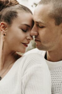 Ein in weiße Pullover gekleidetes Paar teilt einen intimen und liebevollen Moment, der an eine heitere Hochzeit erinnert. Sie stehen sich nah, haben sanft geschlossene Augen und berühren ihre Stirnen. Die Frau trägt Ohrringe und beide wirken zufrieden und in Frieden.