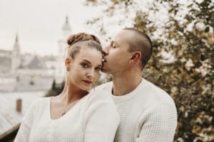 Ein Mann und eine Frau in weißen Pullovern posieren eng beieinander vor dem Hintergrund verschwommenen Bäumen und Gebäuden, was an urbane Romantik erinnert. Der Mann küsst sanft die Stirn der Frau, ein zärtlicher Moment, der ihre Zufriedenheit einfängt und auf zukünftige Pläne für ein Hochzeitsglück hindeutet.