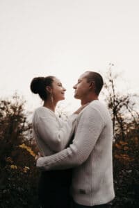 Ein Paar in passenden weißen Pullovern umarmt sich im Freien, umgeben von Herbstbäumen. Die Frau lächelt und sieht den Mann an, der zurückblickt. Die sanfte Atmosphäre schafft eine malerische Kulisse, die an verträumte Hochzeitsplanung unter einem bedeckten Himmel erinnert.