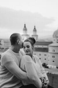 Ein Paar umarmt sich auf einem Dach, der Mann küsst die Stirn der Frau. Sie tragen passende Pullover und fangen einen zeitlosen Moment vor einer Stadtlandschaft mit Kuppeln und Türmen ein. Das Schwarz-Weiß-Bild weckt Romantik und deutet vielleicht auf ihre zukünftige Hochzeitsplanung hin.