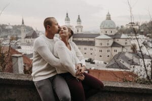 Ein Paar in kuscheligen Pullovern sitzt auf einem Steinsims, während der Mann die Stirn der Frau küsst. Sie blicken auf eine historische Stadtlandschaft mit Kuppeln und Türmen, die sich perfekt für zukünftige Hochzeitsplanungen eignet. Der bewölkte Himmel und das Herbstlaub rahmen die Aussicht wunderschön ein.