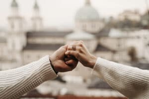 Zwei Menschen in weißen Pullovern stoßen vor einer verschwommenen Stadtlandschaft mit Kuppelgebäuden mit den Fäusten zusammen und symbolisieren damit Einheit und Freude. Eine Person, mit Ringen geschmückt, scheint ihre Hochzeit zu feiern.