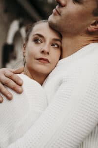 Eine Frau lehnt sich sanft an einen Mann, beide tragen weiße Pullover und verkörpern die Wärme einer Hochzeit. Sie blickt nachdenklich zur Seite, während der Mann sein Kinn auf ihren Kopf legt und einen Moment der Intimität und Liebe einfängt, der sich perfekt für ihre Hochzeitsplanung anfühlt.