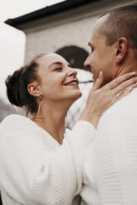 Ein Paar in weißen Pullovern umarmt sich freudig draußen, wobei die Frau den Mann am Hals hält und lächelt, während sie sich liebevoll anschauen und sich ihre bevorstehende Hochzeit vorstellen.