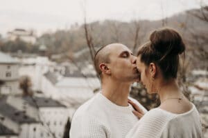 Ein Paar in weißen Pullovern umarmt sich, während der Mann die Frau zärtlich auf die Stirn küsst. Sie stehen im Freien, im Hintergrund eine verschwommene Stadtlandschaft und Hügel – ein perfekter Moment auf ihrer Hochzeitsreise.