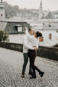 Ein Paar umarmt sich auf einer Kopfsteinpflasterstraße, wo historische Gebäude und Kirchtürme eine romantische und ruhige Kulisse bilden, perfekt zum Heiraten. In gemütlicher, legerer Kleidung scheinen sie in einem Moment verloren zu sein, der so bezaubernd ist wie jede sorgfältig ausgearbeitete Hochzeitsplanung.