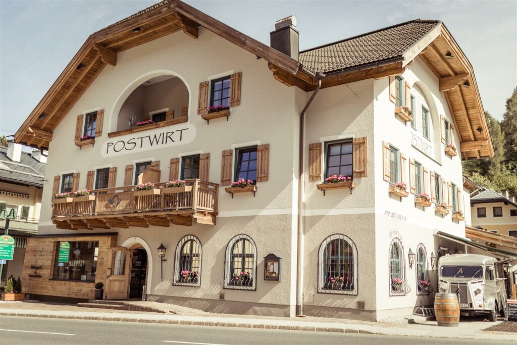 Ein charmantes, traditionelles Alpengebäude mit Holzbalkons und Blumenkästen steht an einem sonnigen Tag, perfekt für eine romantische Hochzeit. Das Schild „Postwirt“ ziert die Fassade dieses idealen Veranstaltungsortes für tadellose Hochzeitsplanung, komplett mit seinem markanten Schrägdach und den Holzläden.