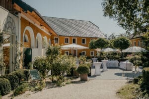 Ein bezaubernder Innenhof mit gelben Gebäuden, gewölbten Fenstern und üppigem Grün erwartet Sie für Ihre Hochzeit. Tische mit weißen Tischdecken und Sonnenschirmen sind für eine elegante Feier aufgestellt. Die Sonne wirft sanfte Schatten und schafft eine warme und einladende Atmosphäre, die perfekt für Hochzeiten ist.