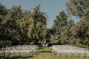 Eine Hochzeit im Freien an einem sonnigen Tag besteht aus zwei Reihen weißer Stühle vor einem schlichten, mit Blumen geschmückten Altar. Üppige grüne Bäume umgeben die ruhige Umgebung und ein paar Leute haben sich in der Nähe des Altars versammelt – perfekt für diejenigen, die ihre Traumhochzeit planen.
