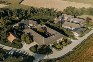 Eine Luftaufnahme eines großen ländlichen Anwesens, perfekt für eine Hochzeit, mit mehreren miteinander verbundenen Gebäuden und einem Innenhof, umgeben von Feldern und Bäumen. Ein paar Autos sind entlang der nahe gelegenen Straße geparkt, während die Landschaft im warmen Sonnenlicht des späten Nachmittags badet.