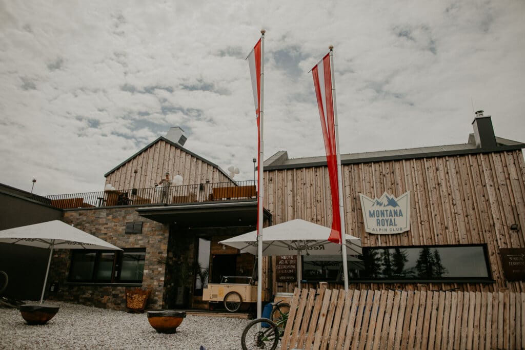 Das Bild zeigt ein rustikales Gebäude mit Holz- und Steinakzenten namens „Montana Royal Alpin Club“. Davor sind zwei Flaggen zu sehen, dazu weiße Sonnenschirme und Sitzgelegenheiten. Der Himmel ist bedeckt und bietet eine romantische Kulisse, die perfekt für die Hochzeitsplanung ist.
