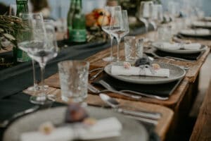Ein rustikaler Esstisch aus Holz ist elegant für ein Hochzeitsessen gedeckt, mit Tellern mit gerollten Servietten, Kristallgläsern und grünen Glasflaschen. Dekoratives Grün und kleine Blumenarrangements unterstreichen das anspruchsvolle Ambiente, perfekt für eine intime Hochzeitsplanungsfeier.