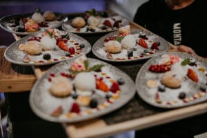 Teller mit Gourmet-Desserts, darunter Kugeln Eis, verschiedene Beeren, Gebäck und Soßen, werden bei einer Hochzeit auf einem Tablett arrangiert, das von einer Person getragen wird. Die Gerichte werden mit Minzblättern garniert und mit Puderzucker bestäubt, was sie perfekt für jede elegante Feier macht.