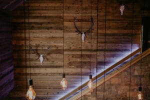 Ein rustikales Interieur, perfekt für eine Hochzeit, mit Holzwänden, die mit montierten Geweihen und Totenköpfen geschmückt sind. Freiliegende Glühbirnen hängen von der Decke und verbreiten ein warmes Licht, das ideal für Hochzeiten ist. Ein beleuchteter diagonaler Holzbalken trennt die Holzwand von einer mit Steinfliesen ausgelegten Wand.