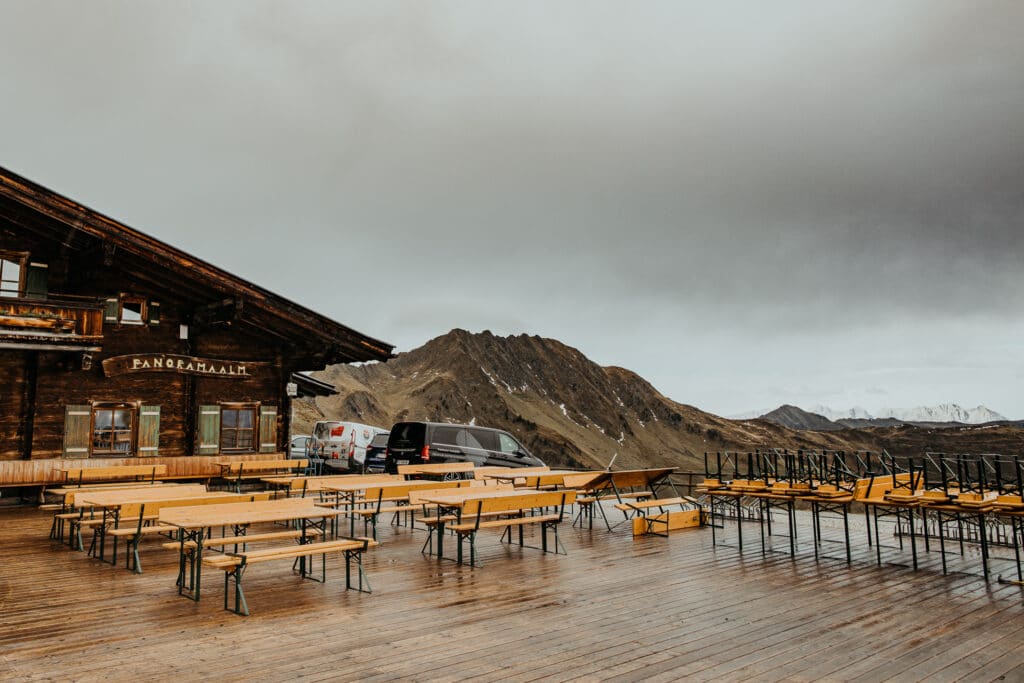 Eine verlassene Außenterrasse mit Holztischen und Bänken liegt vor einer rustikalen Berghütte unter einem bewölkten Himmel, perfekt für intime Momente. Im Hintergrund erheben sich majestätisch die Berge und bieten eine idyllische Kulisse für diejenigen, die von einer unvergesslichen Hochzeit träumen.