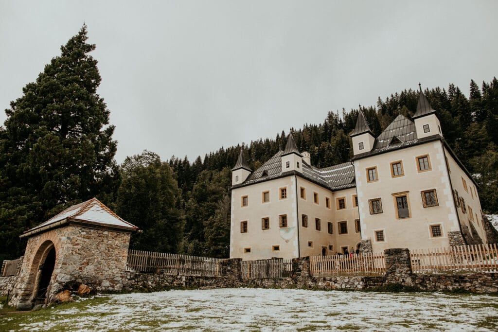 Ein historisches Schloss mit vier spitzen Türmen steht inmitten einer verschneiten Landschaft, perfekt für eine Traumhochzeit. Umgeben von einem Steinzaun und hohen Bäumen ist seine ruhige Atmosphäre ideal zum Heiraten. Ein angrenzendes kleines Steingebäude mit Holzdach vervollständigt diese bezaubernde Kulisse.