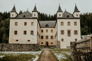 Ein großes historisches Schloss mit vier Türmen steht vor einer bewaldeten Kulisse, perfekt für die Hochzeitsplanung. Der Weg zum Eingang ist von Steinmauern gesäumt und leichter Schnee bedeckt Teile des Grases. Der bewölkte Himmel sorgt für eine stimmungsvolle Atmosphäre, ideal für eine Märchenhochzeit.