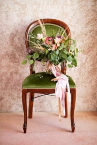 Ein verzierter Holzstuhl mit grüner Polsterung, perfekt für eine Hochzeit, hält einen Blumenstrauß mit rosa und orangefarbenen Blüten, grünem Laub und langen rosa Bändern. Er steht elegant vor einem strukturierten beigen Hintergrund, ideal, um die Essenz der Hochzeitsplanung einzufangen.