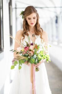 Eine Braut in einem weißen Kleid steht drinnen und hält einen farbenfrohen Blumenstrauß mit rosa und grünen Blumen in den Händen. Ihr langes Haar ist locker gewellt und mit einem kleinen Blumenaccessoire verziert. Sie blickt sanft auf den Blumenstrauß, umgeben von hellem, natürlichem Licht – eine perfekte Szene für die Hochzeitsplanung.