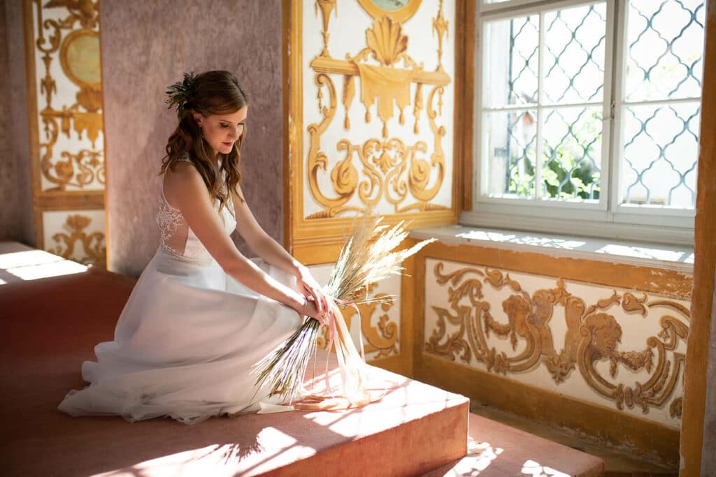Eine Frau in einem weißen Kleid sitzt auf Stufen in einem sonnendurchfluteten Raum mit kunstvollen goldenen Wanddesigns und verkörpert die Gelassenheit einer Hochzeit. Sie hält einen Strauß getrockneter Gräser in der Hand, während Licht durch ein Fenster mit dekorativen Scheiben fällt und diese idyllische Szene der Hochzeitsplanung vervollständigt.