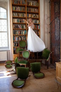 Eine Frau in einem weißen Kleid – das an ein Hochzeitskleid erinnert – steht auf einem Stapel grüner Stühle und greift nach Büchern in einem hohen Bücherregal. Die Holzböden und großen Fenster des Raums tragen zur eleganten, skurrilen Atmosphäre bei, als ob sie sich auf die perfekte Hochzeit vorbereitet.