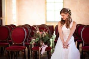 Eine Braut in einem weißen Kleid sitzt neben einem Blumenstrauß auf einem Stuhl in einem leeren Raum mit roten Stühlen und ist in Gedanken über ihre bevorstehende Hochzeit versunken. Mit welligem Haar und Blumenschmuck blickt sie nachdenklich nach unten, während sanftes Tageslicht den Raum erfüllt.