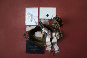Flache Anordnung von Hochzeitspapeterie auf kastanienbrauner Oberfläche. Enthält elegante Einladungskarten, Umschläge mit Wachssiegel, ein dekoratives silbernes Tablett und weiche, gerollte Stoffbänder. Blumenakzente sind kunstvoll neben den Artikeln platziert, perfekt für die Präsentation anspruchsvoller Hochzeitsplanung.