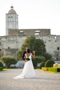 Braut und Bräutigam umarmen sich auf einem Kiesweg vor einem alten Steinschloss, eine zeitlose Szene für ihre Hochzeit. Das lange weiße Kleid der Braut bildet einen Kontrast zum dunklen Anzug des Bräutigams. Grüne Gärten säumen den Weg als Teil ihrer sorgfältigen Hochzeitsplanung, und hinter der Schlossmauer erhebt sich ein Turm.