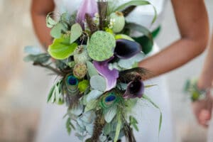 Eine Person hält einen farbenfrohen Blumenstrauß mit violetten Calla-Lilien, grünen Lotusblüten, Eukalyptusblättern und Pfauenfedern, der jeder Hochzeit eine einzigartige Note verleiht. Dieses farbenfrohe Arrangement hebt sich wunderschön von einem sanften, neutralen Hintergrund ab und ist ideal für eine durchdachte Hochzeitsplanung.