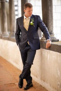 Ein Mann in einem marineblauen Anzug mit hellgoldener Weste und Boutonniere lehnt gedankenverloren an einer Steinsäule. Eine Hand in der Tasche, scheint er über seine bevorstehende Hochzeit nachzudenken, während die sorgfältig kuratierte Hochzeitsplanung in der malerischen Umgebung deutlich wird.