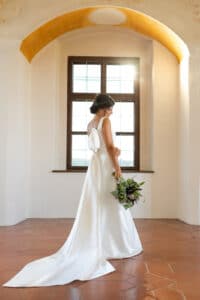 Eine Braut in einem eleganten weißen Kleid steht anmutig da und hält einen Blumenstrauß in der Hand. Während sie den Moment vor ihrer Hochzeit genießt, strömt Sonnenlicht durch ein großes Fenster und beleuchtet die gewölbte Decke und den Terrakottaboden des Raums.